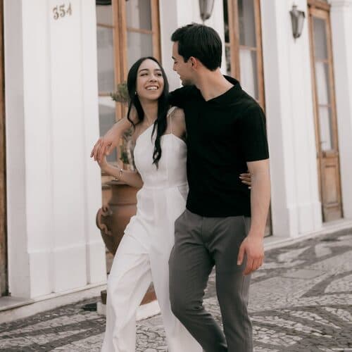 A couple walking arm in arm over calcada cobbles past a white facade